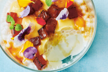 The tropical pannacotta jar by Reynold Poernomo featurees a an overhead shot of a glass bowl willed with white pannacotta, edible flowers and coulis. It is against a light blue background.
